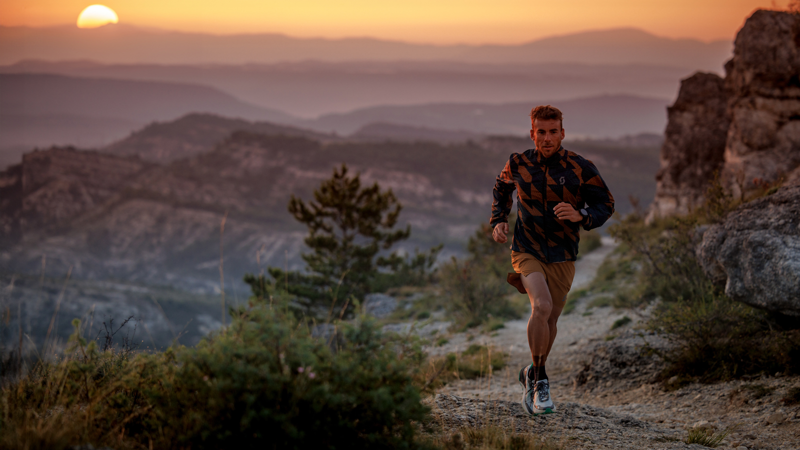 Male trail runner in a patterned jacket and shorts, running on a rugged mountain path at sunset. The scenic background features rolling hills and a golden sky, perfect for outdoor running and adventure sports themes.