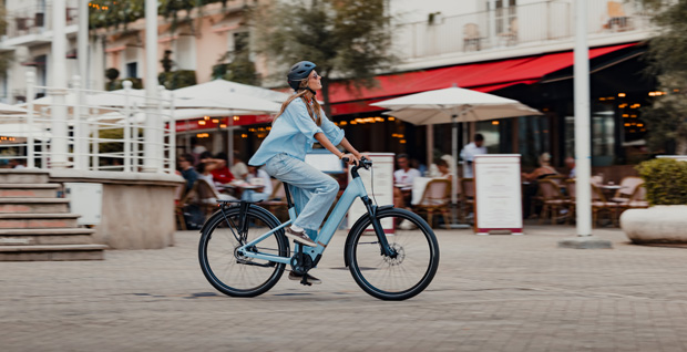 Woman cyclist in casual attire riding a blue electric bike in a bustling city square with outdoor cafes.